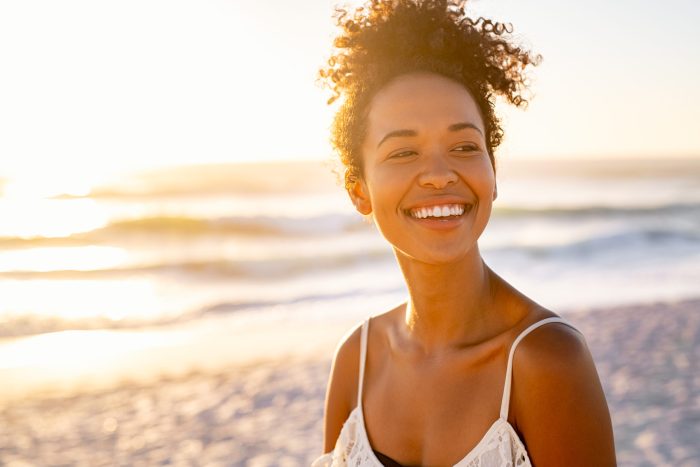 Smiling young black woman in beachwear enjoy sunset at beach. Satisfied beautiful girl with afro hair relaxing at beach during sunrise with copy space. African american woman daydreaming during sunrise.