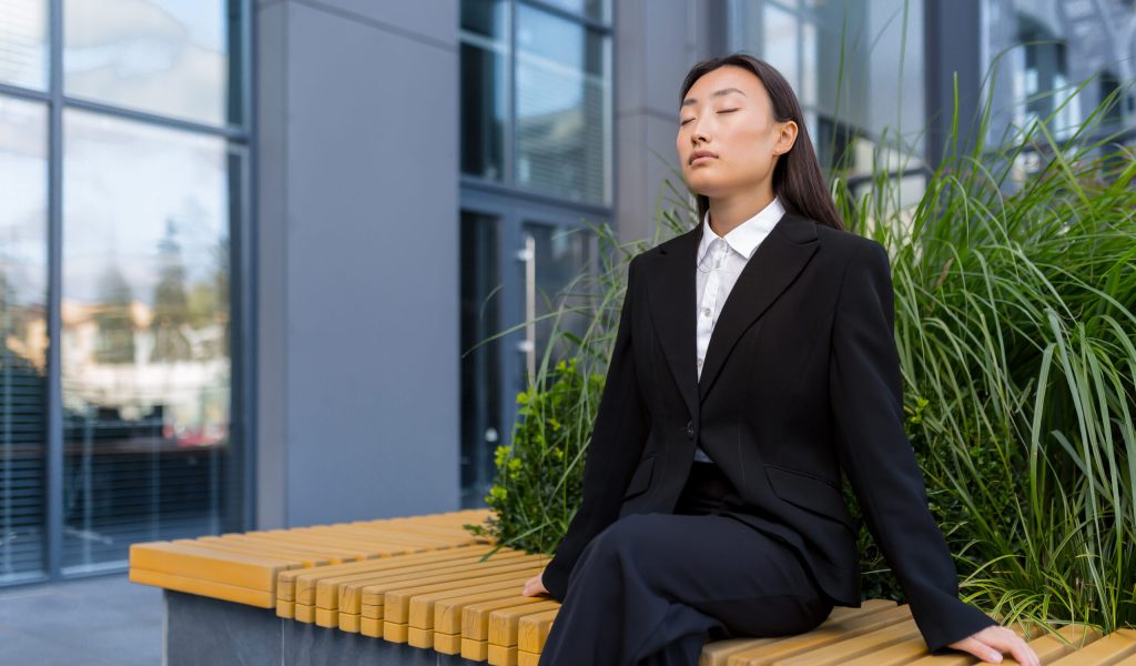 Beautiful Asian business woman sitting on a bench relaxing, meditating and performing breathing exercises
